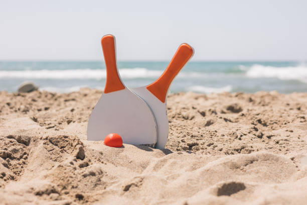 Two rackets and a ball are on the sand, the rackets are orange and white